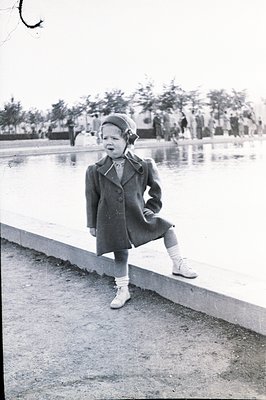A young girl stands confidently beside a reflective pool, wearing a wool coat, knitted hat, and laced-up shoes. Trees and ind...