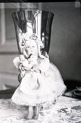 Antique bisque doll posed within a large, ornate glass pedestal vase. The doll wears a ruffled dress and bonnet, seated in a ...