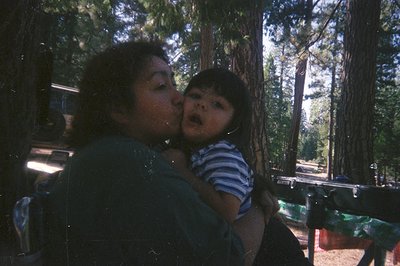 A woman in a dark shirt embraces and kisses a young child held close. The background shows a sunny, forested area suggesting ...