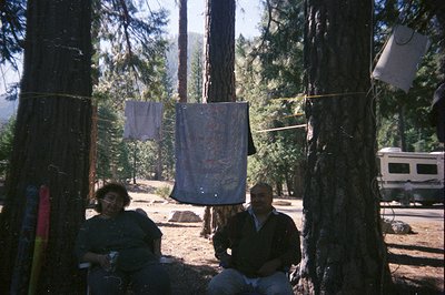 A man and woman seated outdoors near towering pine trees; laundry hangs on a clothesline strung between trees. RV visible in ...
