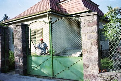 Stone-faced gatehouse with a red tile roof and green-painted metal gate. A person in a red hat trims greenery nearby. Likely ...