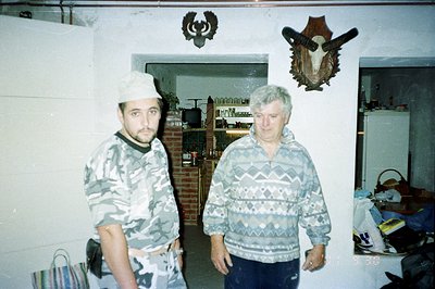 Two men stand in a home interior, one in a camouflage suit and ushanka hat, the other in a patterned sweater. A mounted anima...