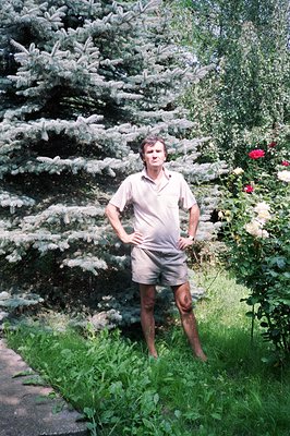 Man in short sleeves and khaki shorts stands with hands on hips, posed before a backdrop of blue spruce trees & flowering ros...