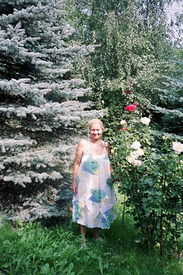A mature woman stands amidst lush greenery, including a prominent, blue-tinted conifer and a rose bush with blooming flowers....