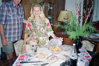 A couple with a dog are posed at a dining table laden with food. Floral table covering, vintage lamps, and wood paneling sugg...