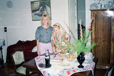 Two women stand near a table laden with fruit and food. The decor, floral tablecloth and framed artwork, suggests a domestic ...