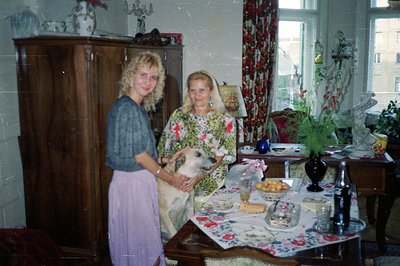 A young woman and elderly woman stand with a dog in a richly decorated dining room. Floral wallpaper and patterned curtains s...