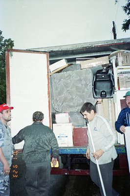 Moving crew loads boxes and furniture into a truck bed. Visible are cardboard boxes, a suitcase, and upholstered items. The t...