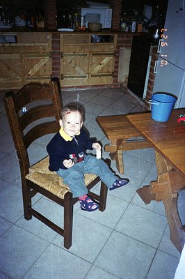 Young boy seated on a woven-seat chair, holding a snack. The kitchen features brickwork, dark wooden cabinetry, and a vintage...