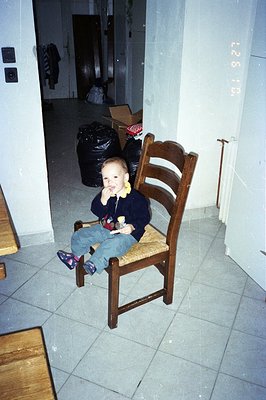 A toddler sits on a woven-seat chair in what appears to be an unfinished home interior. Visible are stacked boxes, black tras...