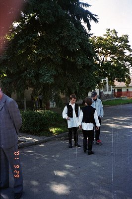 Three boys in traditional folk attire walk along a tree-lined street. The photo captures a candid moment, likely a cultural c...