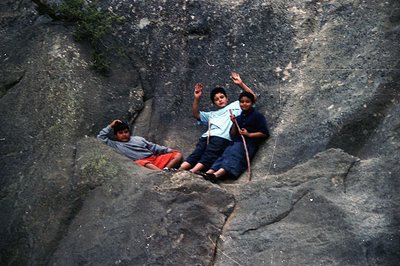 Three boys pose atop a rocky outcrop, likely a natural feature in a mountainous region. They appear to be enjoying a casual m...