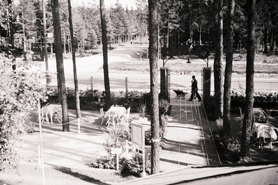 Monochrome view of a park or garden path, featuring a man with a stroller moving towards a decorative archway. Tables & chair...