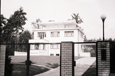Striking modernist villa with flat roof, ribbon windows, and projecting upper level. Brick perimeter wall & gate suggest a pr...
