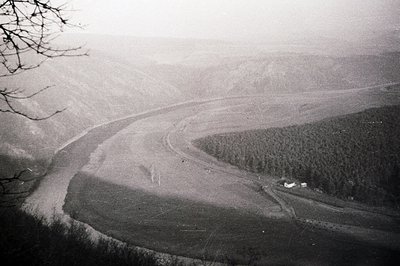 A high-angle view captures a meandering river flowing through a broad, grassy valley. Dense evergreen forest lines the right ...