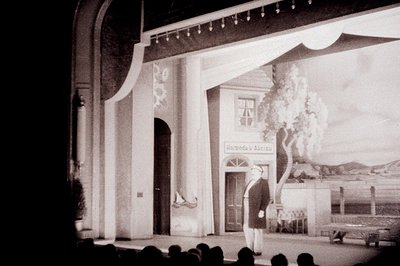 Stage backdrop depicts a seaside building facade and landscape, likely representing a coastal town. Cyrillic script visible o...