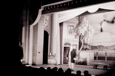 Stage backdrop depicts a Mediterranean-style building with arched doorway and signage (likely Cyrillic). A male performer in ...