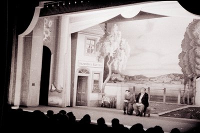 Stage backdrop depicts a European storefront & rural scene; a man and woman sit in chairs. The backdrop’s text, “Raspoda u Al...