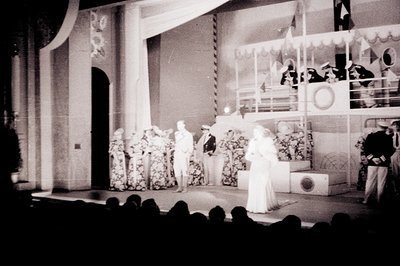 A theatrical stage scene, possibly a musical or revue. A woman in a long gown stands center stage, facing a group of men in n...