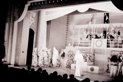 Stage backdrop depicts a seaside scene with a ship and balcony. Performers in period costumes are posed on a raised platform....