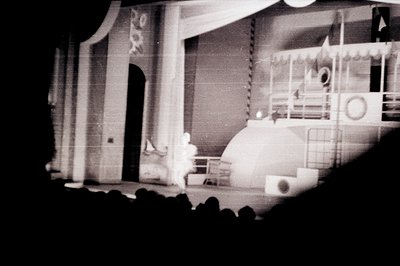 A staged scene, likely from a theatrical production or film set. Features a stylized, modernist architectural backdrop with a...