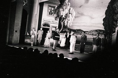 A staged scene depicts a woman in a long dress, a uniformed officer, and other figures near a painted backdrop featuring a bu...