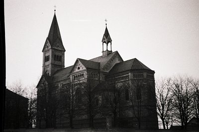 A grayscale photograph captures a stately church building with a steeply pitched roof, a central tower, and a smaller flankin...