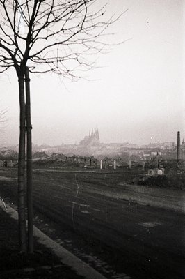 A hazy, monochrome view captures a distant cityscape dominated by a cathedral's spires. A foreground road leads towards the u...