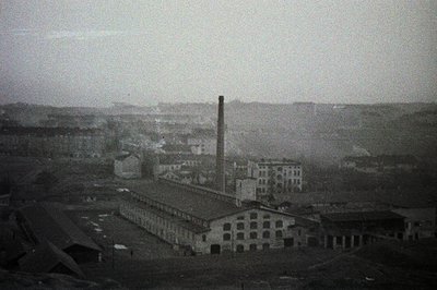 Industrial complex, likely a factory or mill, dominates the view with a tall smokestack. Multi-story buildings with numerous ...
