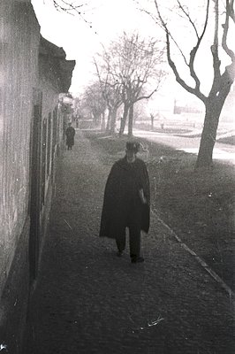 A solitary figure in a long coat and hat walks along a tree-lined path. A weathered wall defines the left side of the frame. ...