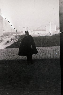 A formally dressed man in a dark cape and hat stands on a brick-paved surface, viewed from behind. A large, industrial constr...