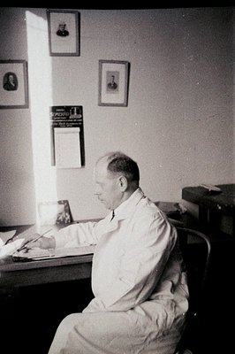 A man in a lab coat sits at a table with papers, seemingly concentrating on his work. Three framed prints adorn the wall. The...