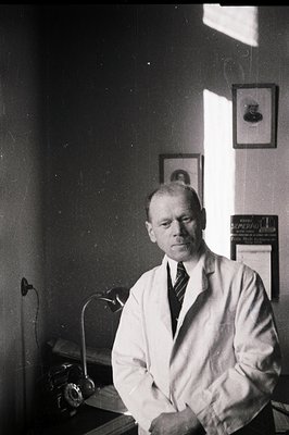 A formally posed portrait of a man in a white lab coat, seated behind a desk. He wears a dark tie and appears to be in a doct...