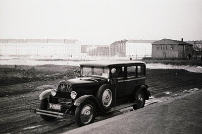 A black & white image of a 1930s sedan, likely a Ford, captured on a dirt road. The vehicle bears a license plate "P-25020". ...