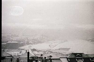 A sweeping, snow-covered landscape with rooftops of buildings in the foreground. A coastal vista stretches towards the horizo...