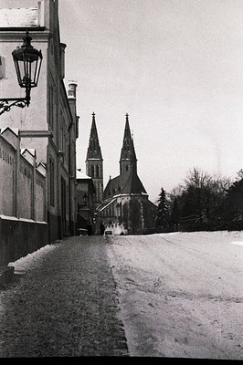 Monochrome view of a cobblestone street ascending toward a grand cathedral with twin spires. A wrought-iron lamppost and ston...