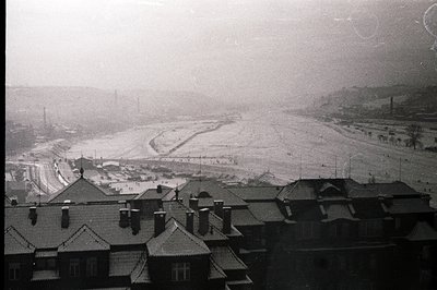 Overlooking a snow-covered, industrial landscape with factory smokestacks. Rooftops of brick buildings dominate the foregroun...