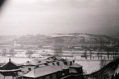 A snowy, overcast landscape reveals a town nestled in a valley, framed by rolling hills. Visible are rooftops with dark, tile...