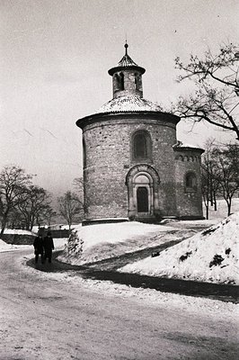 A circular brick structure, likely a chapel or mausoleum, stands amidst a snowy landscape. Two figures are seen approaching a...