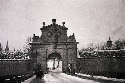 A stark monochrome image captures a monumental stone gateway flanked by a fortified wall, likely a castle entrance. Snow blan...