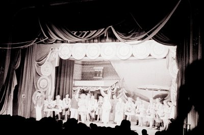 A theatrical stage scene depicts a large group of formally-dressed individuals gathered in front of a painted backdrop of a b...