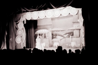A stage production featuring a backdrop depicting a seaside scene with a pier and sailboats. Two figures, likely performers, ...