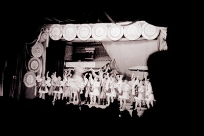 A staged scene depicting a crowded rooftop gathering, likely a theatrical presentation or promotional display. Detailed archi...