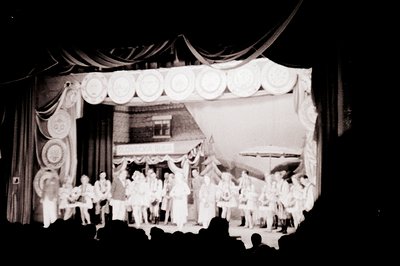 A theatrical stage scene depicts a village market facade with numerous figures—likely actors—dressed in traditional attire. D...
