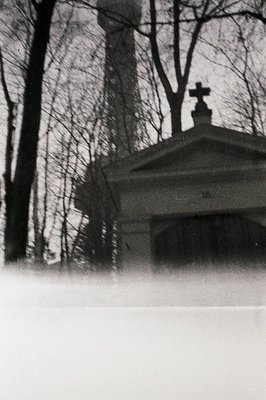 A grayscale image depicts a historic church facade partially obscured by leafless trees and snow. Architectural details are v...