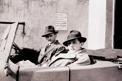 Two men in fedoras and trench coats, seated in an open vehicle, appear to be in motion. A sign written in a foreign language ...