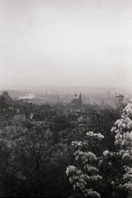 A hazy, atmospheric view of a European cityscape, likely Prague. A prominent church spire rises above the rooftops, partially...