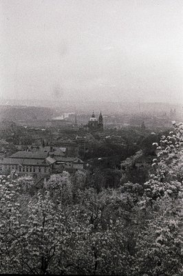 A misty, elevated view of Prague's cityscape. Buildings with distinctive roofs are prominent, with a church dome rising above...