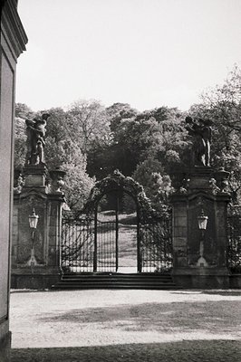 Ornate wrought iron gate flanked by sculpted pillars & lanterns, opening to a manicured lawn & distant foliage. Formal garden...
