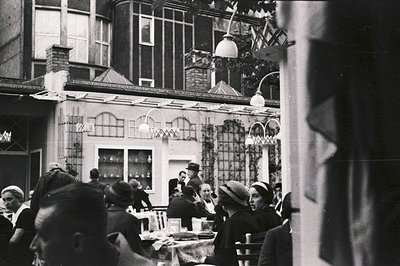 Patrons seated at tables under a pergola roofed with climbing vines. A server in a hat attends to a group. Architectural styl...